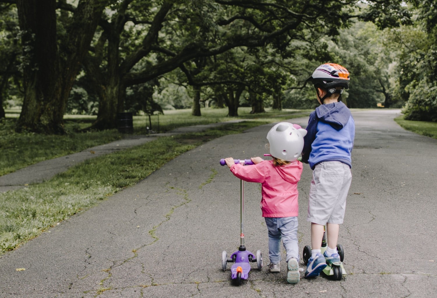 Two children wearing helmets and riding scooters together on a tree-lined park path